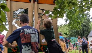 A child and an adult sit on wooden stools at outdoor game stations under trees, playing a hands-on activity at a lively park event with more people and green grass in the background.