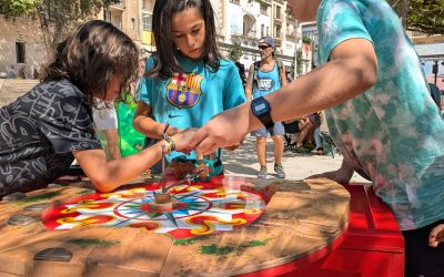 Three children play a colourful spinning top game outdoors on a sunny day, focused on the wooden table top. Other people and buildings appear in the background.