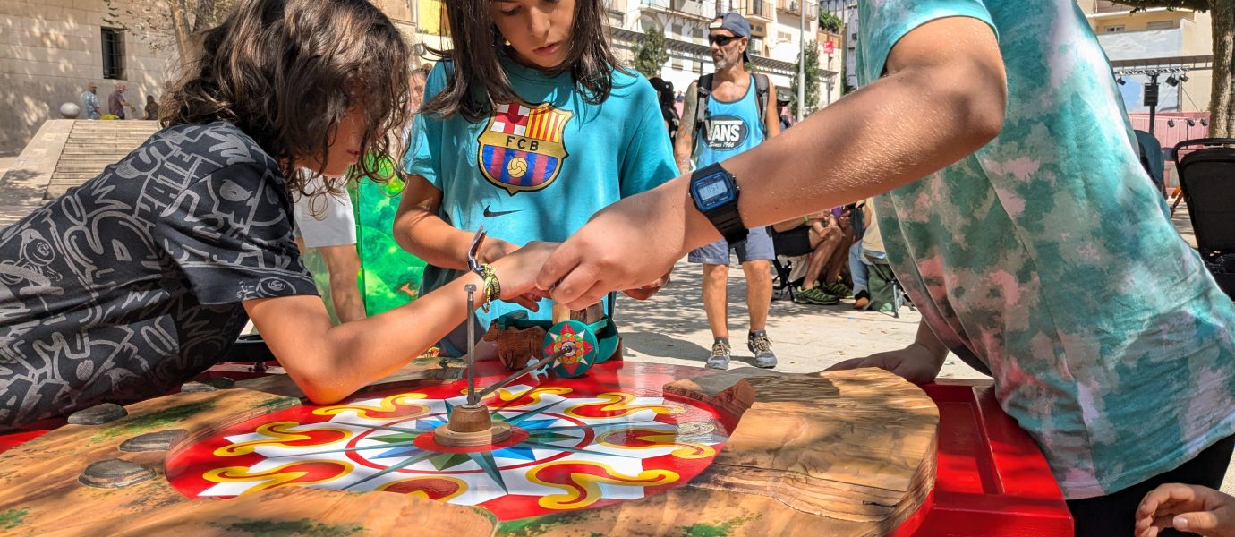 Three children play an outdoor spinning top game on a colourful circular wooden table, focusing intently. Other people and buildings are visible in the sunny background.