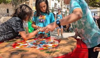Three children play an outdoor spinning top game on a colourful circular wooden table, focusing intently. Other people and buildings are visible in the sunny background.