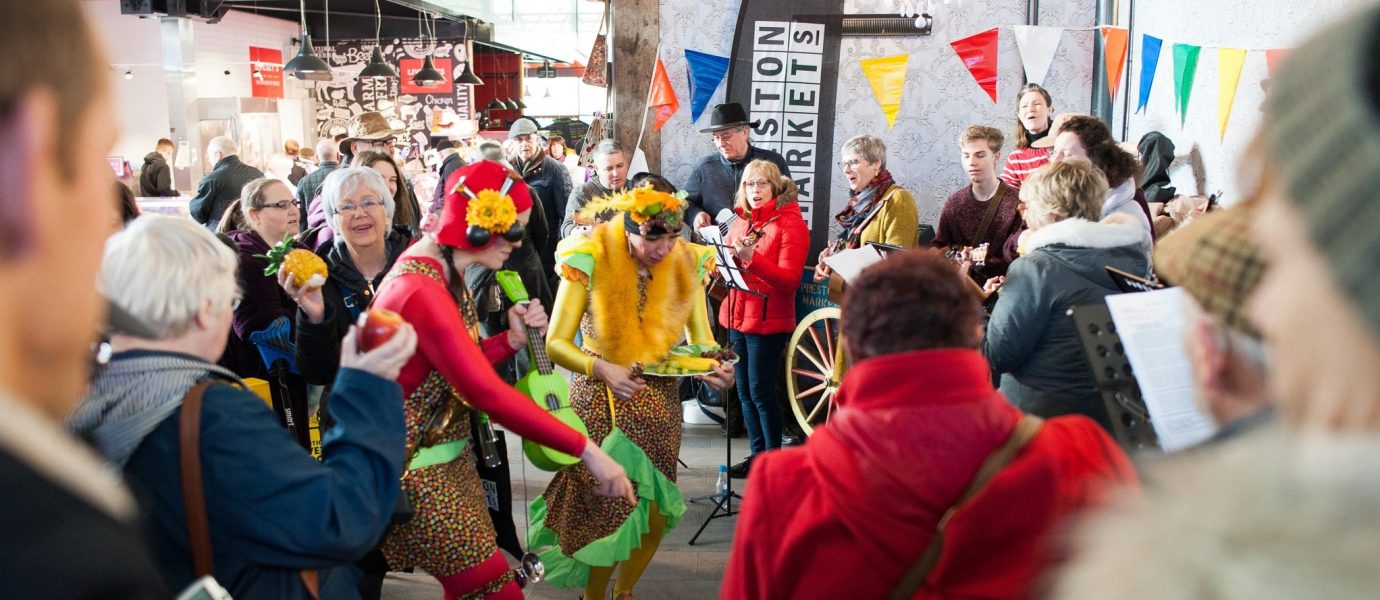 A lively crowd gathers indoors as two performers in colourful, flower-themed costumes dance and play music. People watch and take photos, with market stalls and bunting visible in the background.
