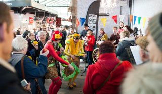 A lively crowd gathers indoors as two performers in colourful, flower-themed costumes dance and play music. People watch and take photos, with market stalls and bunting visible in the background.