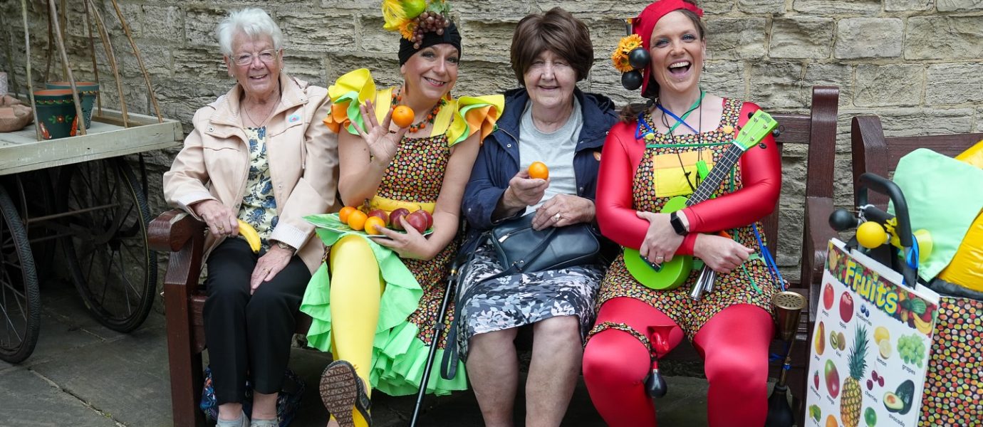 Four women sit on a bench; two wear colourful clown costumes and hold fruit props, while the other two wear casual clothes and smile. A stone wall and a fruit-themed trolley are in the background.
