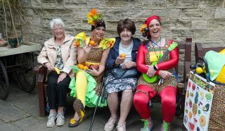 Four women sit on a bench; two wear colourful clown costumes and hold fruit props, while the other two wear casual clothes and smile. A stone wall and a fruit-themed trolley are in the background.