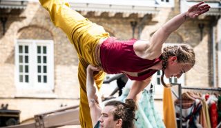 A male performer balances a female acrobat on his hand. She extends one leg and arm gracefully in the air, wearing yellow trousers and a red top. Old brick buildings are visible in the background.