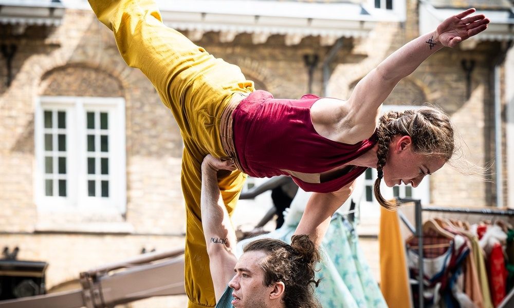 A male performer lifts a female acrobat with one hand during an outdoor show. The woman balances horizontally with one leg extended, wearing yellow trousers and a red top. Historic buildings are visible in the background.