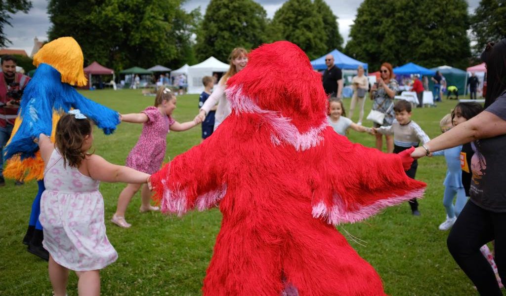 Children and adults hold hands in a circle with two people in colourful, furry costumes (one red, one yellow and blue) in a grassy park. Tents and trees are visible in the background.