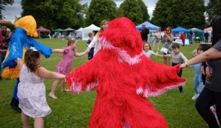 Children and adults hold hands in a circle with two people in colourful, furry costumes (one red, one yellow and blue) in a grassy park. Tents and trees are visible in the background.