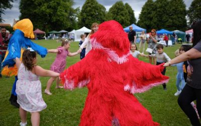 Children and adults hold hands in a circle with two people in colourful, furry costumes (one red, one yellow and blue) in a grassy park. Tents and trees are visible in the background.