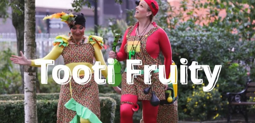 Two performers in colourful, patterned costumes and fruit-themed accessories walk outdoors in a park, smiling. Bold white text across the image reads Tooti Fruity. Trees and bushes are in the background.