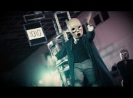 A person wearing a pale, expressionless mask, long coat, and gloves performs on stage with another masked figure in the background; a scoreboard displaying 1010 is visible behind them.