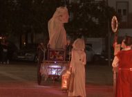 A nighttime outdoor scene shows people in historical costumes. One person on stilts with a draped white figure stands on a cart, while others in white dresses and a person in red with a top hat stand nearby. Spectators watch in the background.