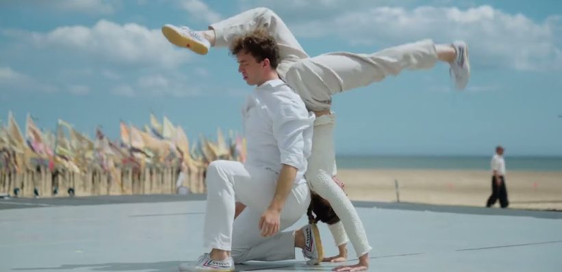Two people dressed in white perform an acrobatic dance on an outdoor stage near a beach, with one person balancing on their hands and the other kneeling. Colourful flags are visible in the background under a partly cloudy sky.