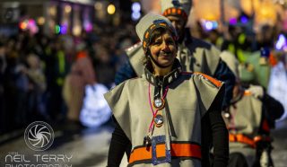 A smiling person in a futuristic grey costume with orange accents walks in a night-time parade, surrounded by others in similar outfits. Bright lights and a crowd are visible in the background.