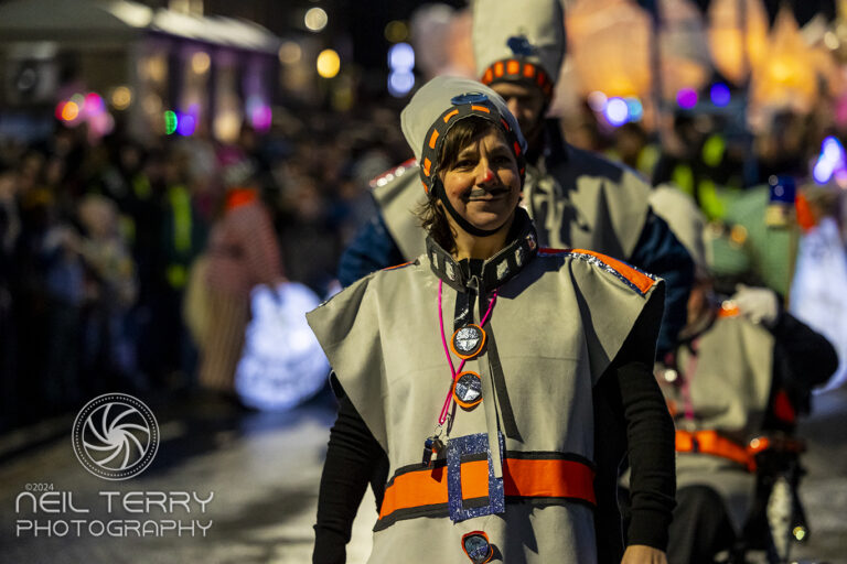 A smiling person in a futuristic grey costume with orange accents walks in a night-time parade, surrounded by others in similar outfits. Bright lights and a crowd are visible in the background.