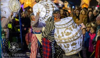 Performers in colourful costumes carry large, illuminated lanterns shaped like barrels, entertaining a crowd of people, including children, during a nighttime street parade or festival.