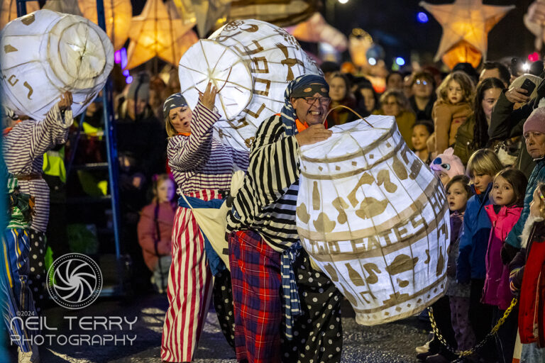 Performers in colourful costumes carry large, illuminated lanterns shaped like barrels, entertaining a crowd of people, including children, during a nighttime street parade or festival.