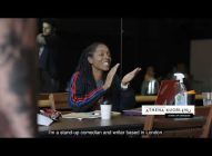 A woman with long plaited hair, wearing a striped jacket, sits at a table with a laptop and notebooks, smiling and gesturing with her hands. Text on screen identifies her as Athena Kugblenu, a stand-up comedian.