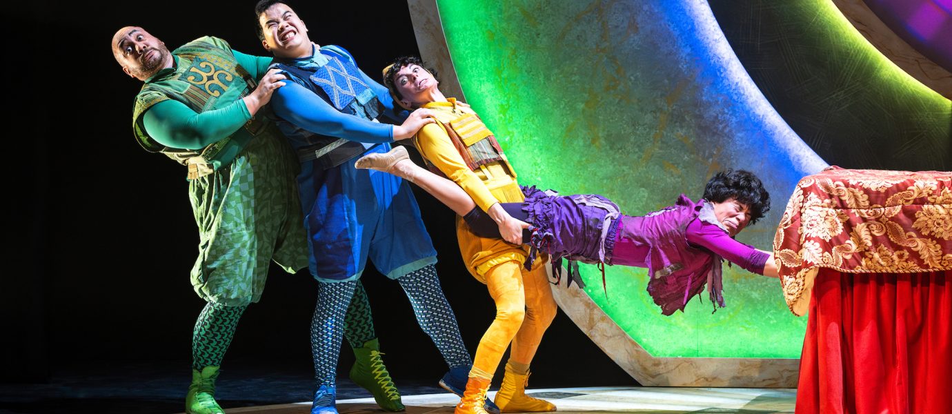 Four actors in colourful, whimsical costumes comically pull each other in a chain on stage. The last person, dressed in purple, holds onto a table covered with a red cloth, while the others are dressed in green, blue, and yellow.