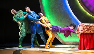 Four actors in colourful, whimsical costumes comically pull each other in a chain on stage. The last person, dressed in purple, holds onto a table covered with a red cloth, while the others are dressed in green, blue, and yellow.