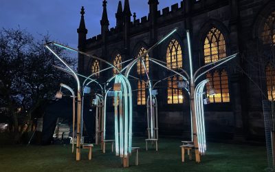 A nighttime outdoor scene shows glowing light sculptures shaped like trees in front of a historic building with stained glass windows, illuminating the grass and benches below.