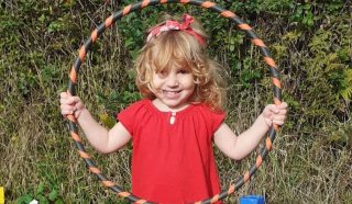 Smiling young girl with curly blonde hair, wearing a red dress and headband, holds a striped hula hoop in front of her whilst standing outdoors in front of green foliage.