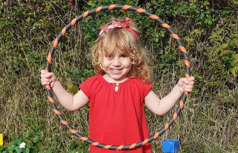 Smiling young girl with curly blonde hair, wearing a red dress and headband, holds a striped hula hoop in front of her whilst standing outdoors in front of green foliage.