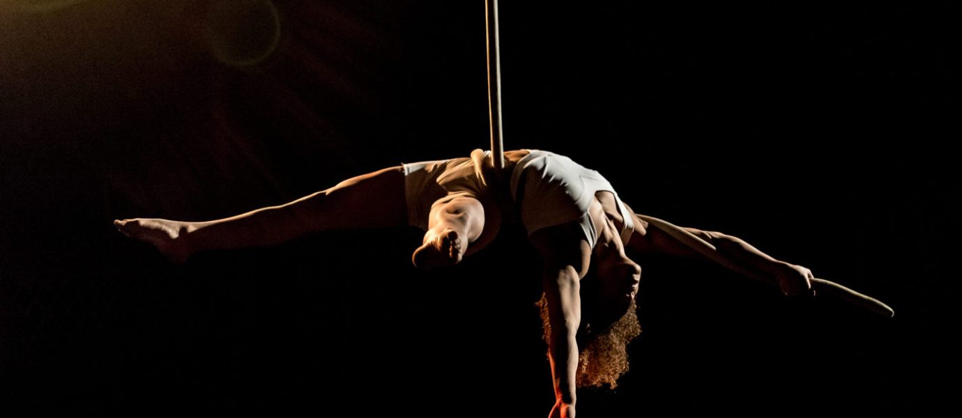 Aerial performer in a white outfit hangs upside down with arms and legs extended on a vertical pole, set against a dark background with dramatic stage lighting.