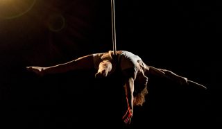 Aerial performer in a white outfit hangs upside down with arms and legs extended on a vertical pole, set against a dark background with dramatic stage lighting.