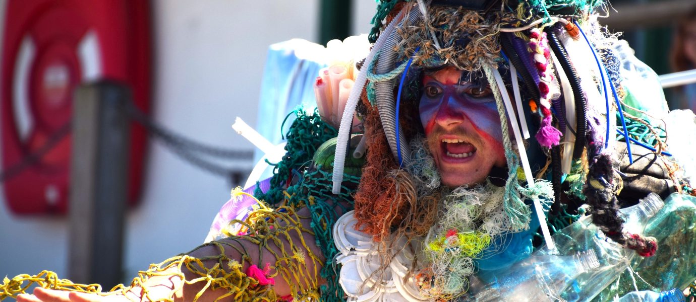 A person in colourful face paint and a costume made of plastic waste, netting, and debris gestures with an outstretched arm, possibly performing or protesting environmental pollution.