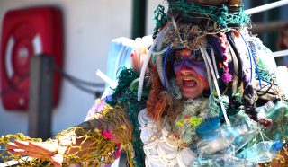 A person in colourful face paint and a costume made of plastic waste, netting, and debris gestures with an outstretched arm, possibly performing or protesting environmental pollution.