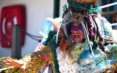 A person wearing a colourful, dramatic costume made of plastic waste and nets, with their face painted boldly, extends their arm and shouts passionately. A red fire hose reel is visible in the background.