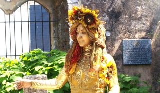 A person dressed as a golden statue, adorned with sunflowers and ornate golden clothing, stands outdoors near a stone wall and green plants, with a plaque visible in the background.