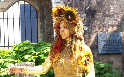 A person dressed as a golden statue, adorned with sunflowers and ornate golden clothing, stands outdoors near a stone wall and green plants, with a plaque visible in the background.