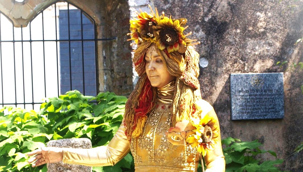 A person dressed as a golden statue, adorned with sunflowers and ornate golden clothing, stands outdoors near a stone wall and green plants, with a plaque visible in the background.