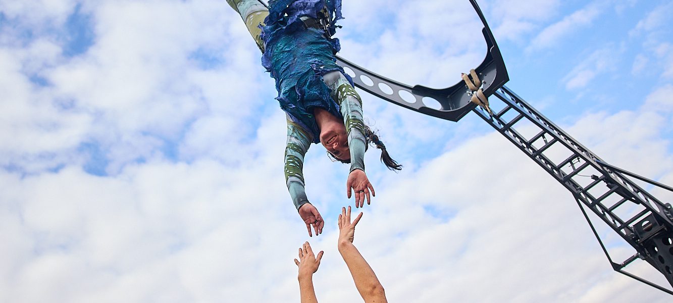 A street performer hangs upside down from a metal frame, reaching towards another performer standing below with arms raised, as a crowd watches in an outdoor urban setting under a partly cloudy sky.