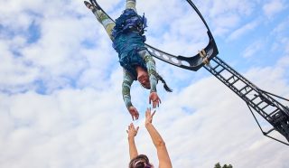 A street performer hangs upside down from a metal frame, reaching towards another performer standing below with arms raised, as a crowd watches in an outdoor urban setting under a partly cloudy sky.
