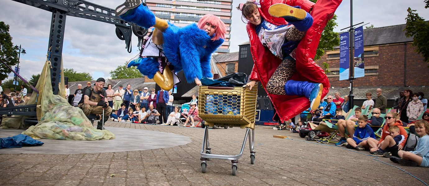 Two performers in colourful costumes, one blue and one red, leap dramatically over a shopping trolley in an outdoor public space, while a crowd of people watches the action. Trees and buildings are visible in the background.