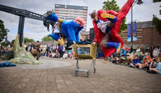 Two performers in colourful costumes, one blue and one red, leap dramatically over a shopping trolley in an outdoor public space, while a crowd of people watches the action. Trees and buildings are visible in the background.