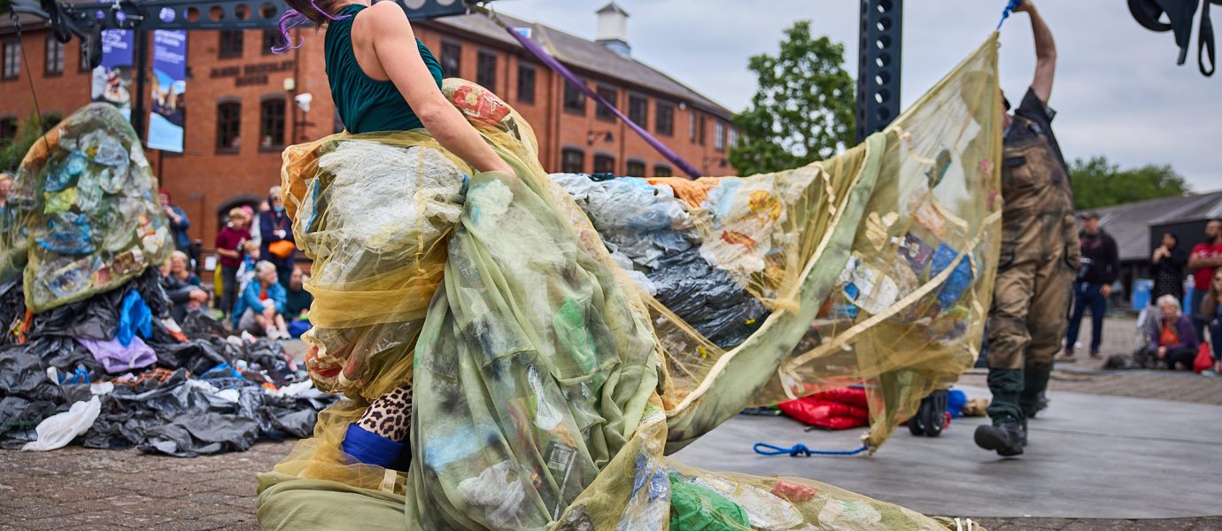 A performer outdoors wears a flowing gown made of netting filled with plastic rubbish, dramatically arching backwards during a street performance, with a crowd and brick buildings in the background.
