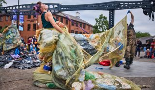 A performer outdoors wears a flowing gown made of netting filled with plastic rubbish, dramatically arching backwards during a street performance, with a crowd and brick buildings in the background.