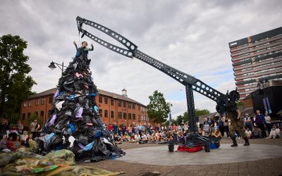 A performer stands atop a large pile of clothes, raised by a mechanical crane, while a crowd watches the outdoor show in an urban square with brick and modern buildings in the background.