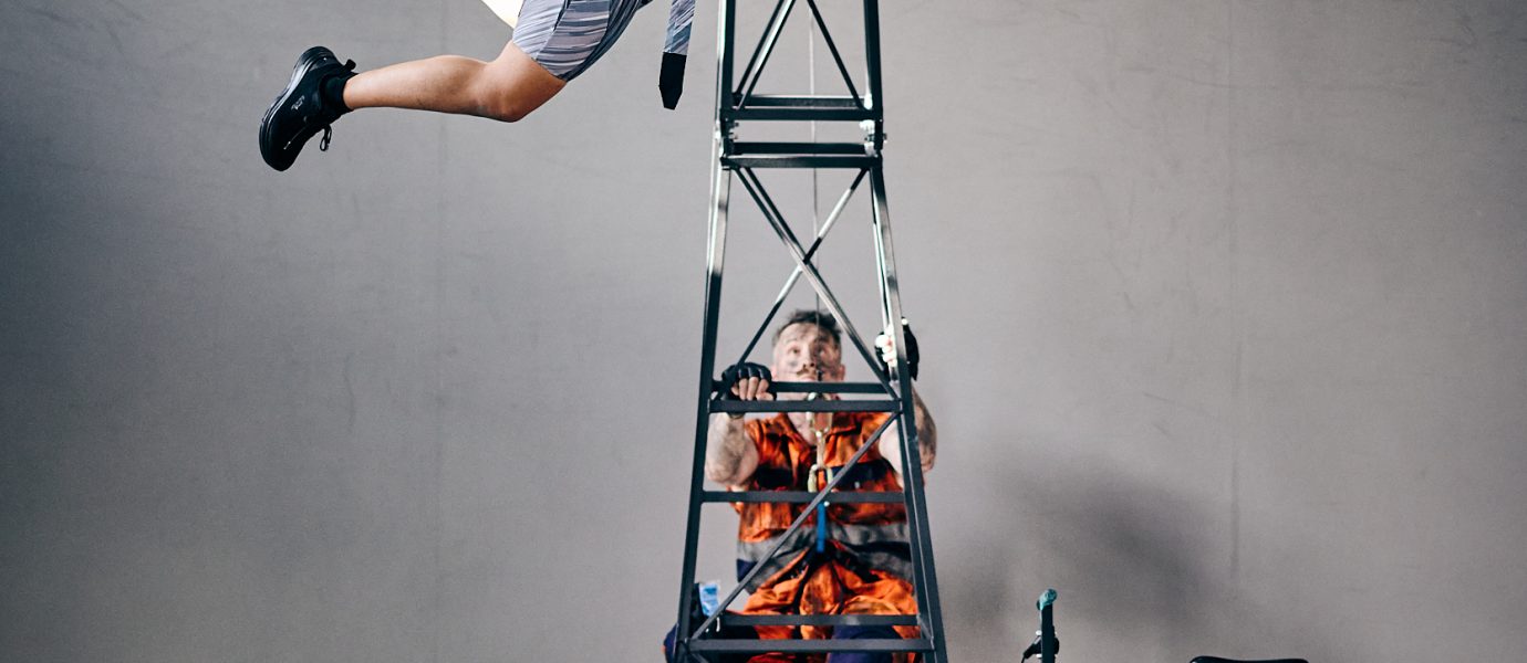 A performer in a striped dress is suspended in mid-air, arms outstretched, attached to a metal tower. Another person in orange sits below, looking up. The scene is industrial and dramatic.