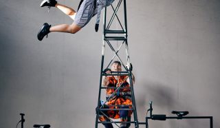 A performer in a striped dress is suspended in the air by a harness attached to a metal structure, arms outstretched. Another person in check sits below, working with ropes and equipment. The scene is indoors.