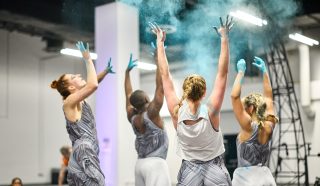 Five dancers in matching outfits throw blue powder into the air while performing indoors, with their backs mostly to the camera and arms raised, the scene vibrant and energetic.