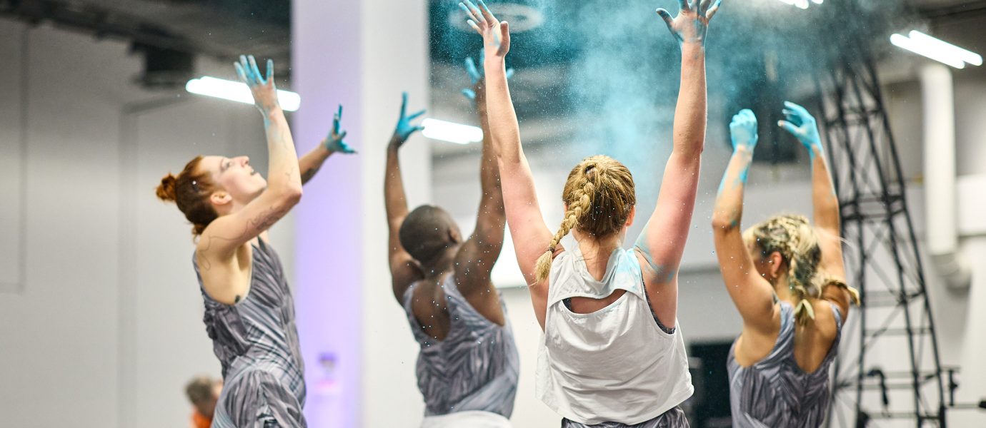 Five dancers in matching grey outfits throw blue powder into the air, their arms raised. The performance takes place indoors, with people and structural columns visible in the background.
