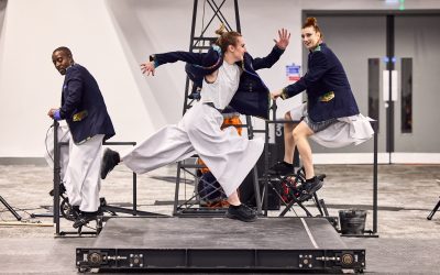 Performers in stylish jackets and wide trousers are captured mid-motion on a platform, one looking forward with excitement, another in mid-stride.