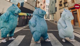 Three people in blue hippopotamus costumes cross an empty city street at a pedestrian crossing, with buildings and recycling bins in the background.