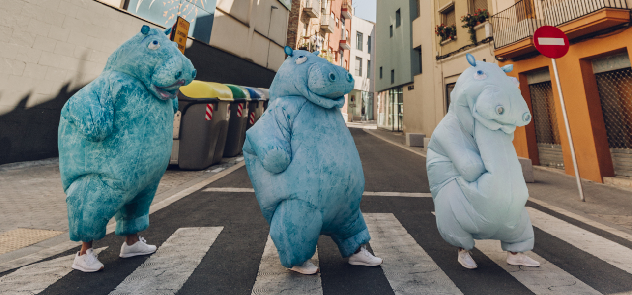 Three people in blue hippopotamus costumes cross an empty city street at a pedestrian crossing, with buildings and recycling bins in the background.