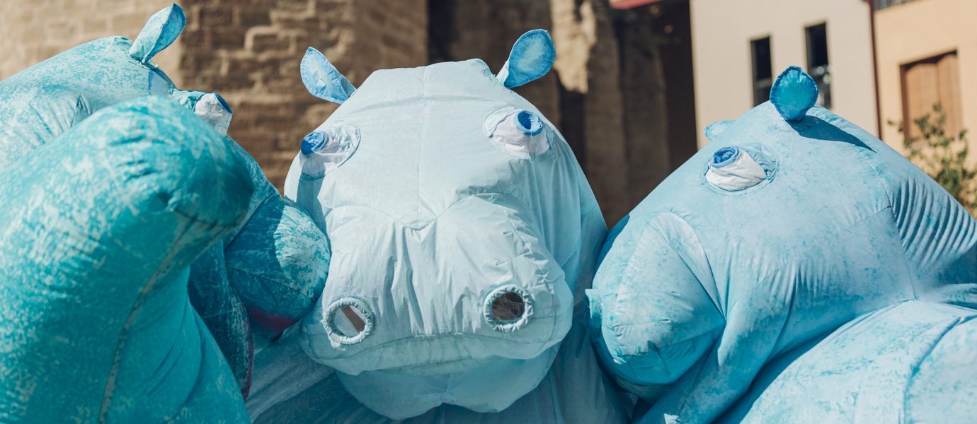 Three people wearing large, blue hippo costumes stand close together outdoors, with buildings and a stone wall in the background. The costumes feature exaggerated hippo faces and nostrils.